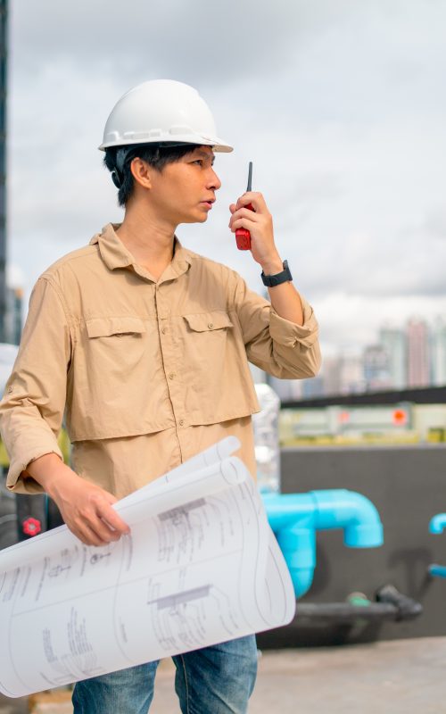 Asian engineer or technician worker with holding blueprint use walkie-talkie to contact with coworker to continue work in construction site and he stay on terrace of building.