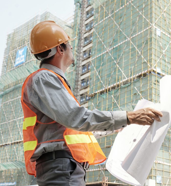 Engineer wearing hardhat and reflective vest looking at high building under construction while standing with blueprint outdoors and checking the sketches